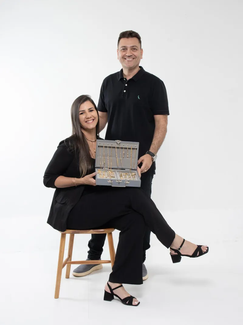 Os donos da marca, de roupa preta e sorrindo: ela sentada num banquinho de madeira, segurando a maleta aberta com colares e anéis dourados, e ele em pé atrás dela, em fundo branco