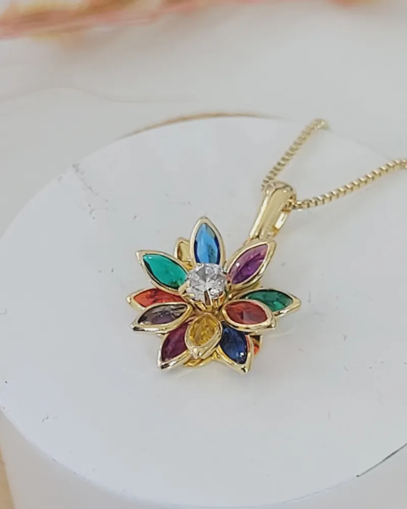 Colar dourado com pingente de flor 3D de pétalas coloridas em cores fortes, azul, verde, roxo, vermelho e amarelo, sobre uma base branca redonda