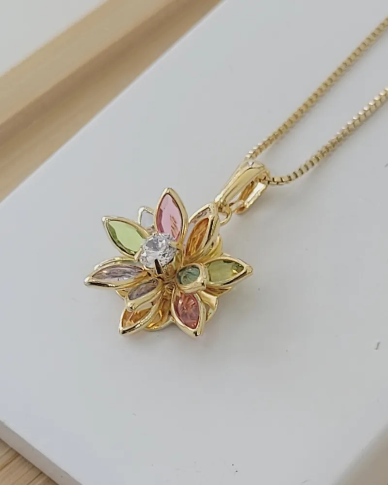 Colar dourado com pingente de flor 3D de pétalas coloridas em cores suaves, rosa, verde, lilás e laranja, sobre uma base branca
