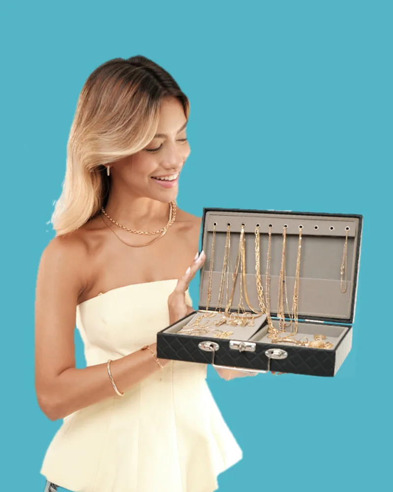 Modelo sorrindo, de top claro, segurando uma maleta preta aberta cheia de colares e pulseiras dourados, em fundo azul