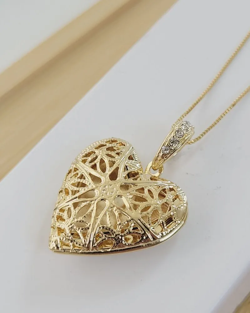 Colar dourado com pingente relicário de coração grande, vazado com desenhos de flores e pedrinhas no passador, sobre uma base branca